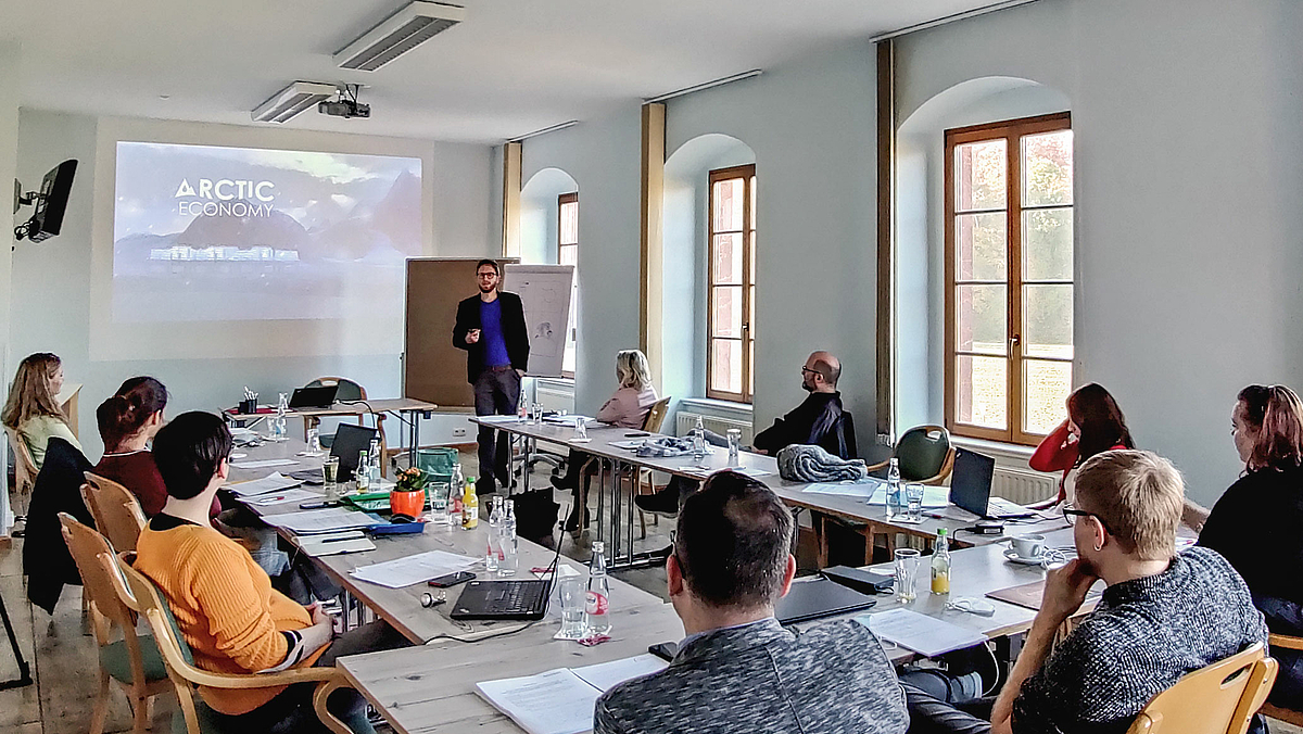 Das Foto zeigt 9 Personen sitzend an Seminartischen. Sie folgen der Präsentation eines jungen Mannes . Auf dem Beamerbild hinter ihm ist eine Grafik mit dem Schriftzug "Arctic Ecomomy" zu lesen.