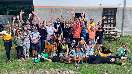Im gemeinsamen Jubel mit den Kindern kaum zu entdecken: Betreuer:innen des Outdoor-Technik-Camps 2023 der Hochschule Mittweida. (Foto: Markus Lohse) Gruppenbild auf einer Wiese vor einem Gebäude mit rund 30 Personen, darunter überwiegend Kinder zwischen 8 und 12 Jahren. Die meisten stehen jubelnd mit nach oben gestreckten Armen, andere sitzen auf dem Boden, einige liegen.