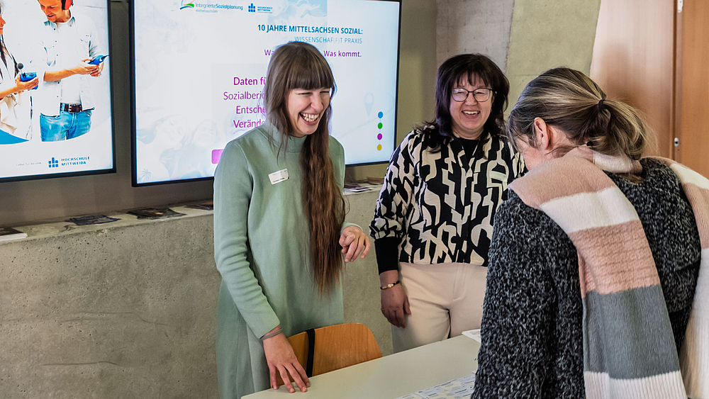 Zwei weibliche Personen freundlich lachend eine Teilnehmerin begrüßend. Hinter ihnen ein Monitor mit „Mittelsachsen Sozial“-Motiv.
