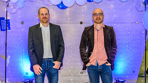 Exzellente Lehre und Betreuung: Florian Heinke (l.) und Professor Alexander Marbach sind ausgezeichnet! Das Foto zeigt zwei männliche Personen vor einer mit weißen und blauen Luftballons sowie Lichterketten geschmückten Wand.