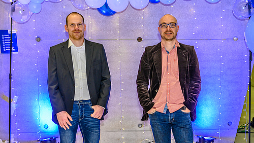 Exzellente Lehre und Betreuung: Florian Heinke (l.) und Professor Alexander Marbach sind ausgezeichnet! Das Foto zeigt zwei männliche Personen vor einer mit weißen und blauen Luftballons sowie Lichterketten geschmückten Wand.