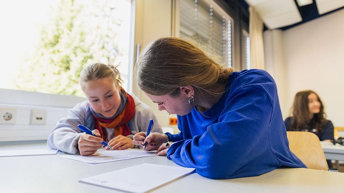 Zwei Mädchen mit Kugelschreibern über einem Schreibblock gebeugt an einem Tisch sitzend