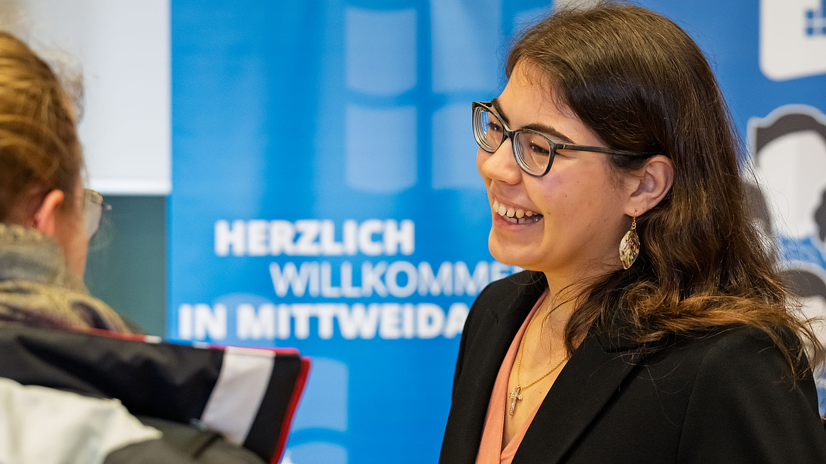 Herzlich lachende junge Frau im Gespräch. In Hintergrund ist auf einem Rollup "Herzlich willkommen in Mittweida" zu lesen.